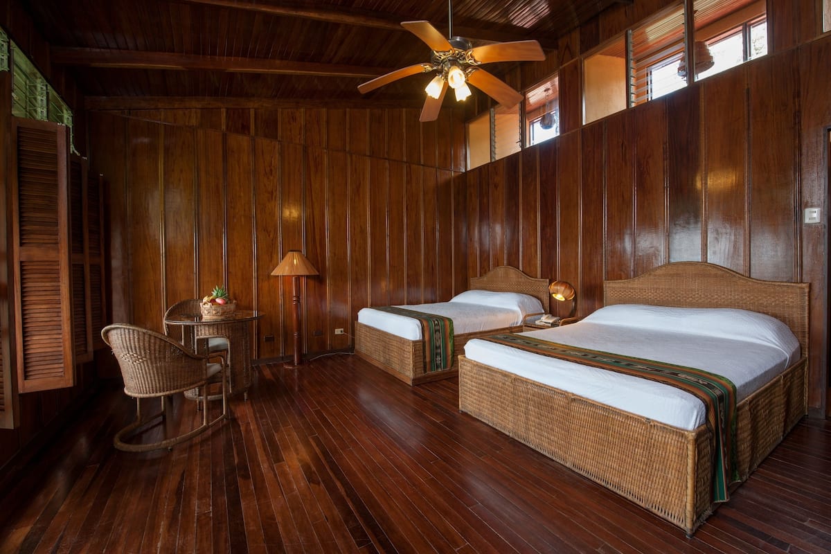 A wooden-paneled room with two single beds, a small table with a lamp, and a wicker chair.