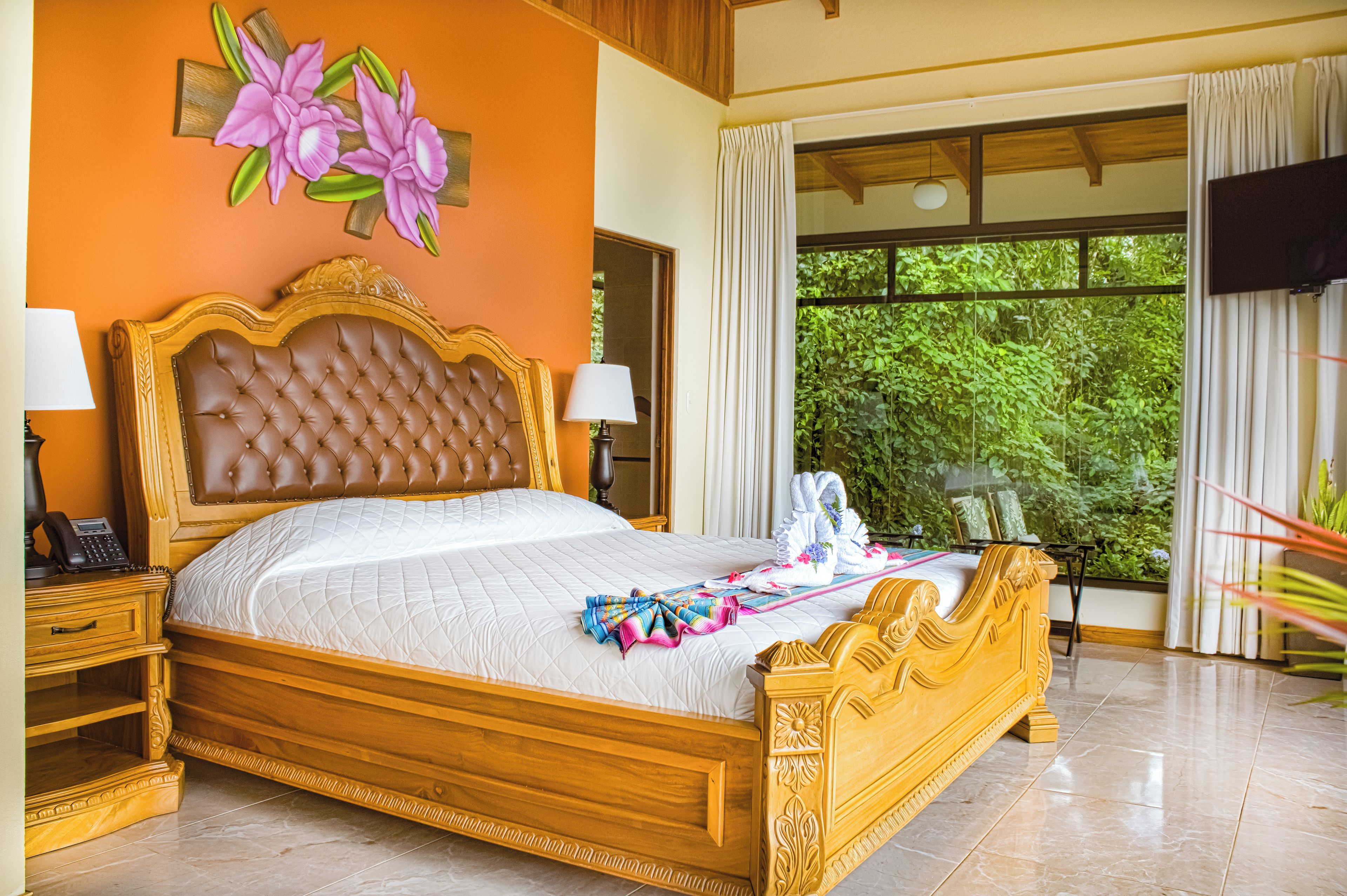 A large bed with a wooden headboard, a bedside table with a lamp, and a view of greenery through the window.
