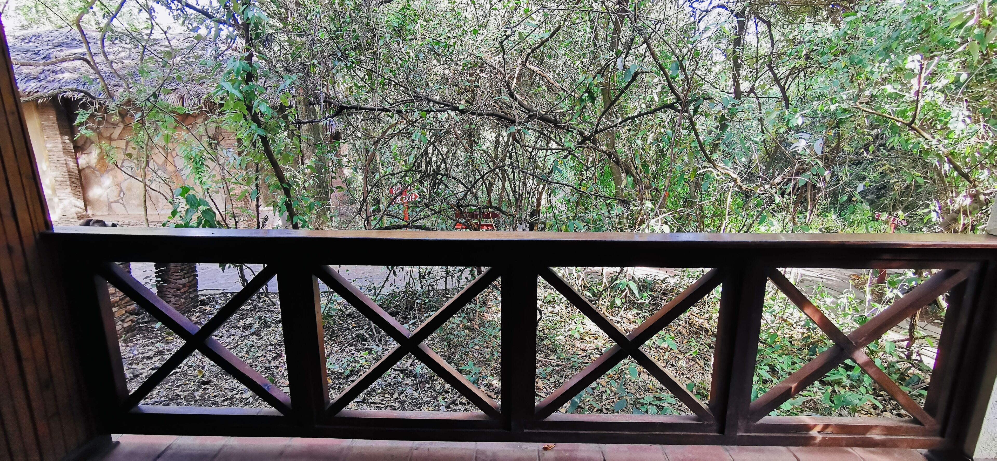 A wooden balcony with a metal railing overlooking a garden area with trees and a stone wall.