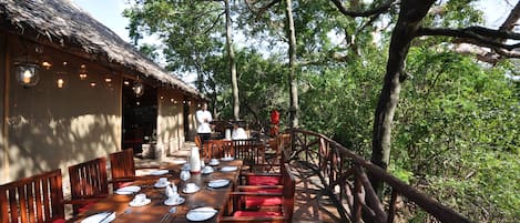 An outdoor dining area with wooden tables and chairs, surrounded by trees and a thatched-roof building.