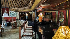 A bar area with wooden chairs and tables, a stone wall, and a thatched roof.