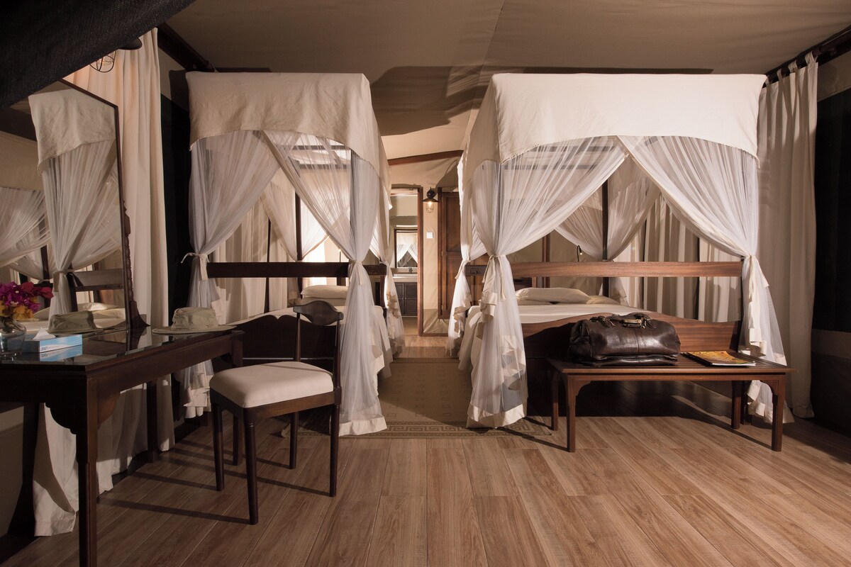 A room with a wooden floor, a desk with a chair, a bedside table with a handbag, and a bed with white curtains.