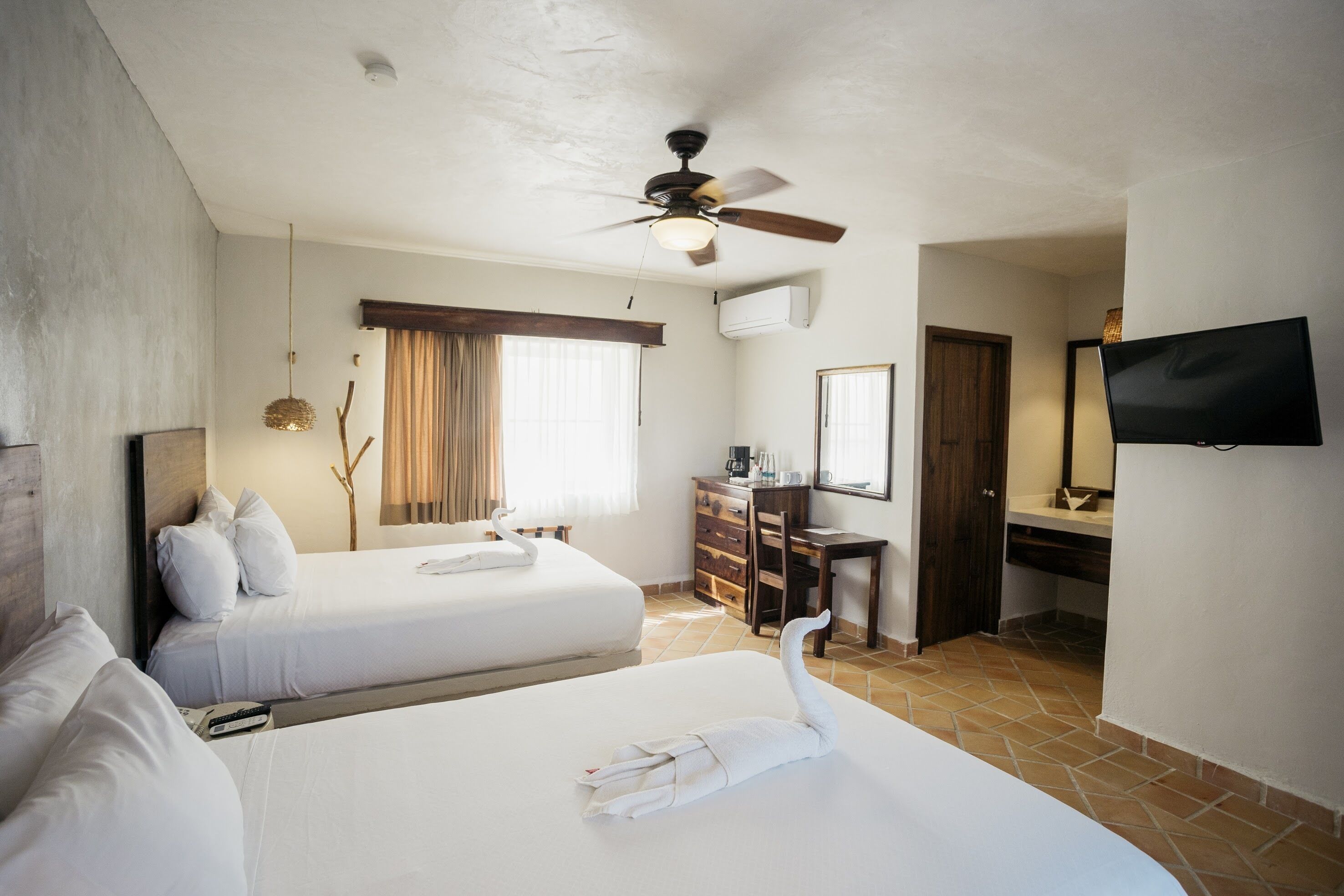 Habitación de hotel con dos camas, ventilador de techo, televisión, escritorio y baño.