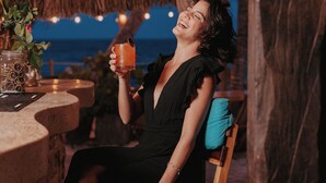A person sitting on a wooden chair with a teal cushion, holding a drink. The setting includes a rustic wooden ceiling and a view of the ocean in the background. - Petit Lafitte Beachfront Hotel & Bungalows (Playa del Carmen)