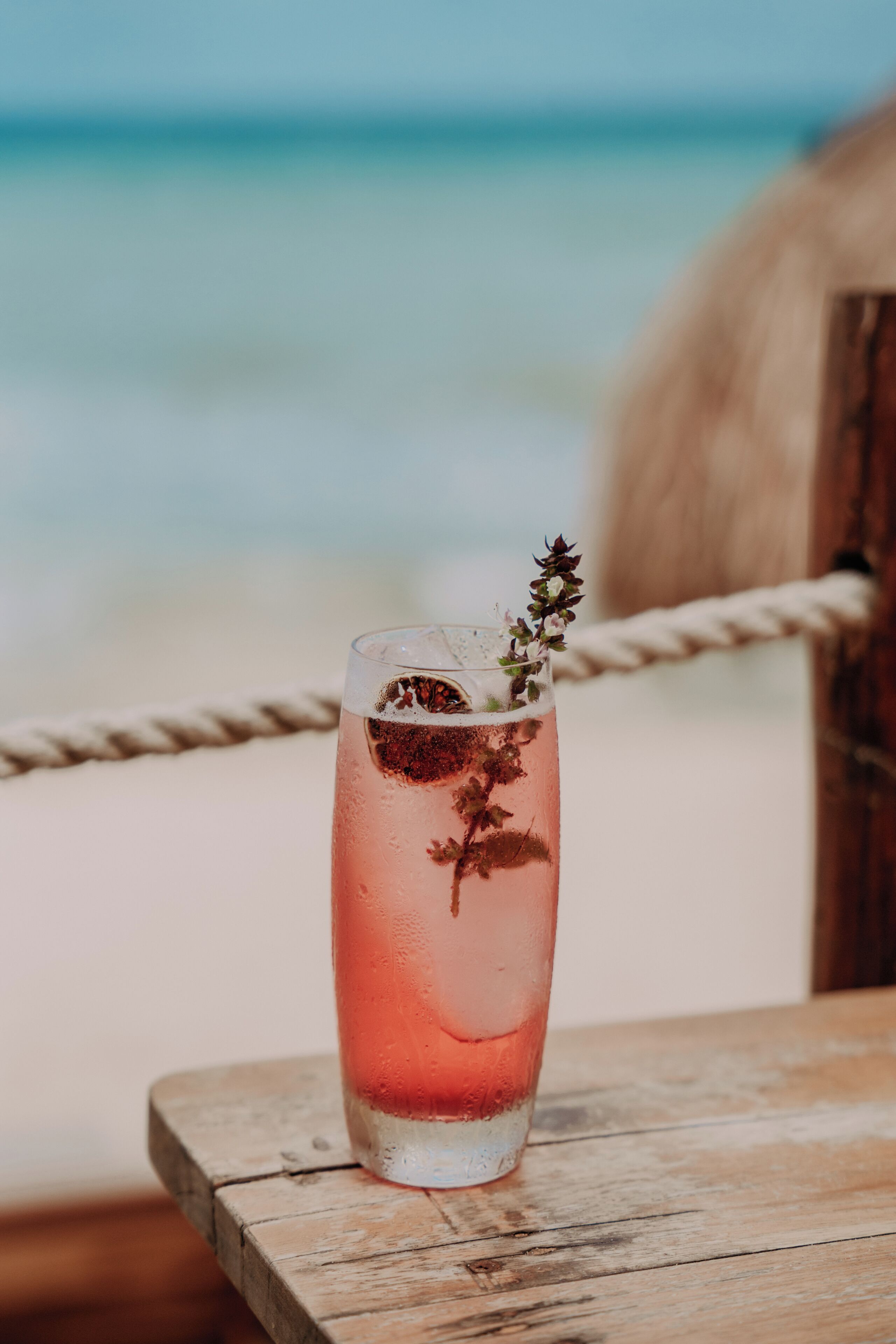 Un vaso de bebida rosada con una ramita de hierba sobre una mesa de madera cerca de la playa.
