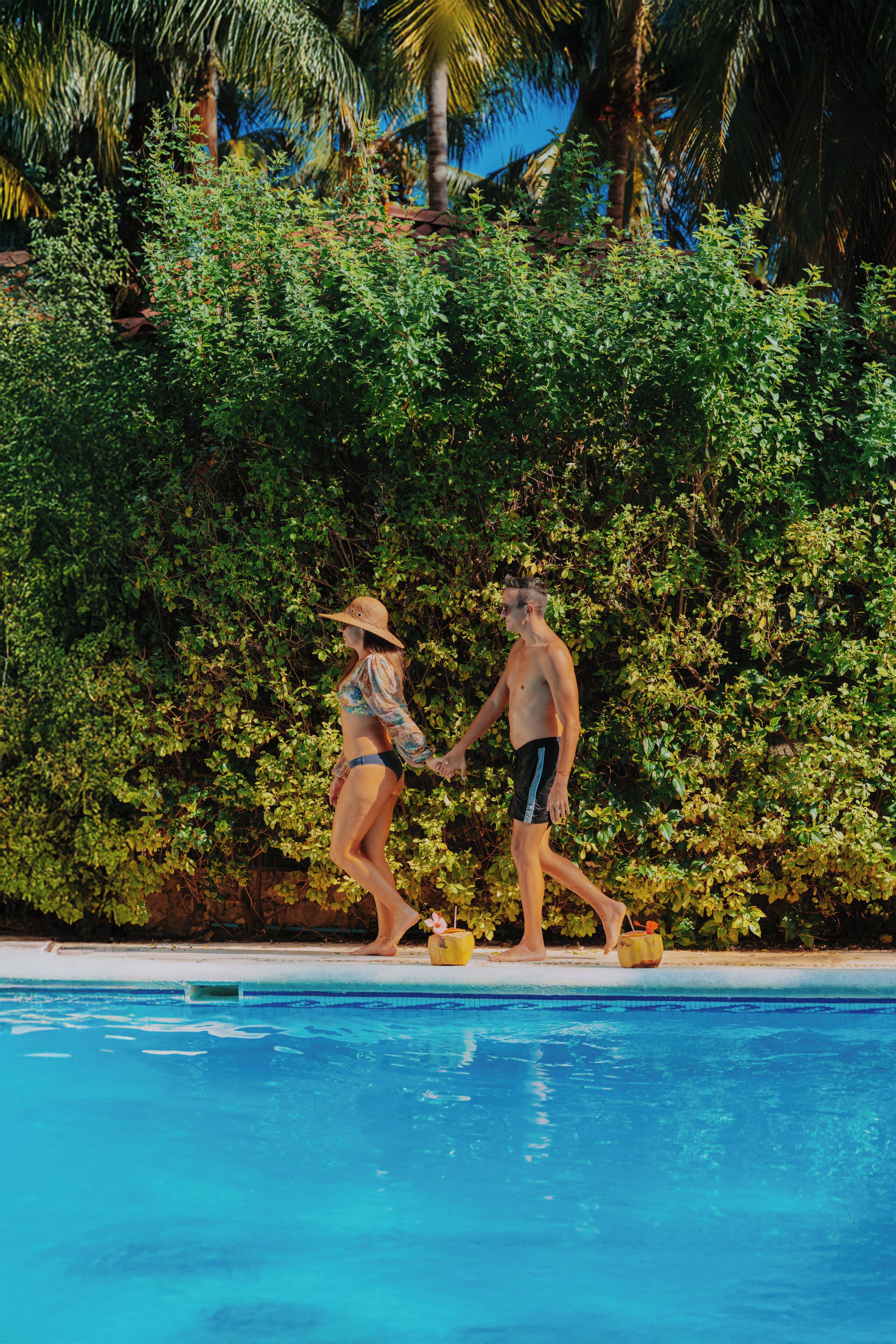 Una pareja caminando junto a una piscina con abundante vegetación y palmeras al fondo.