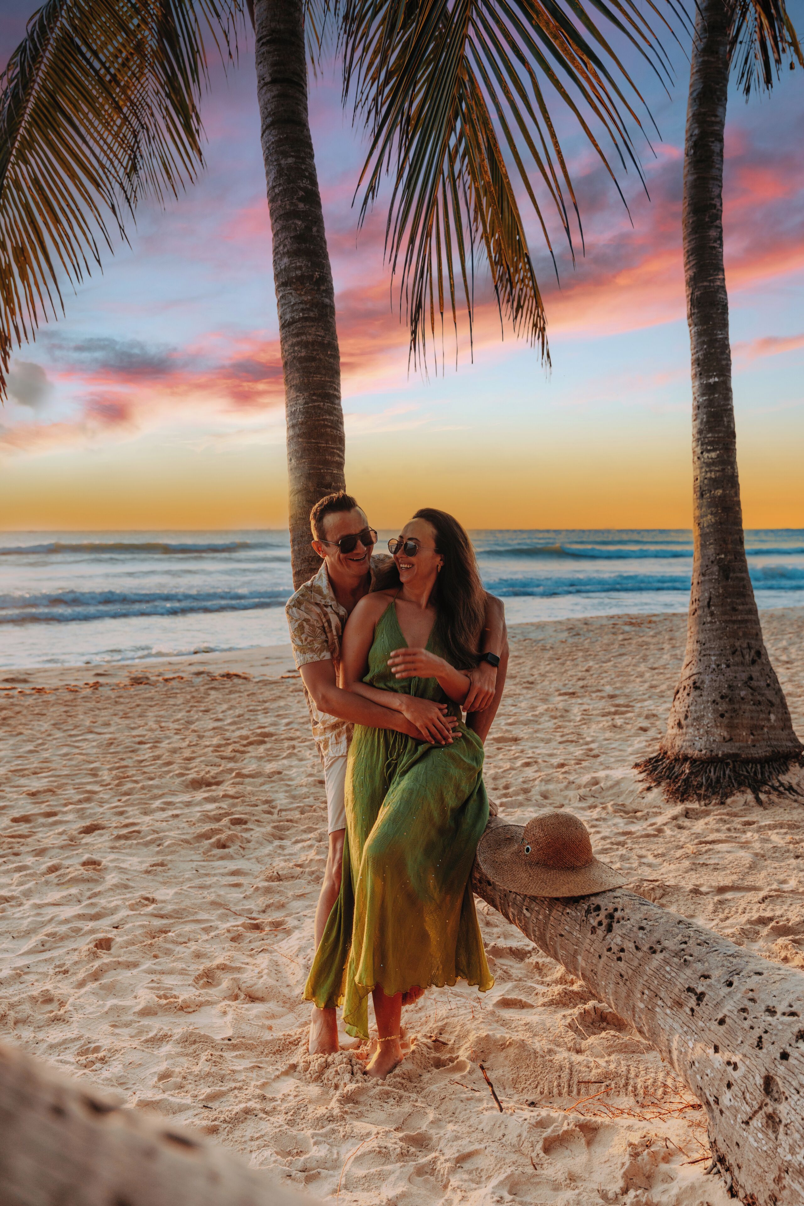 Una pareja se abraza en una playa arenosa, con palmeras y un atardecer de fondo.