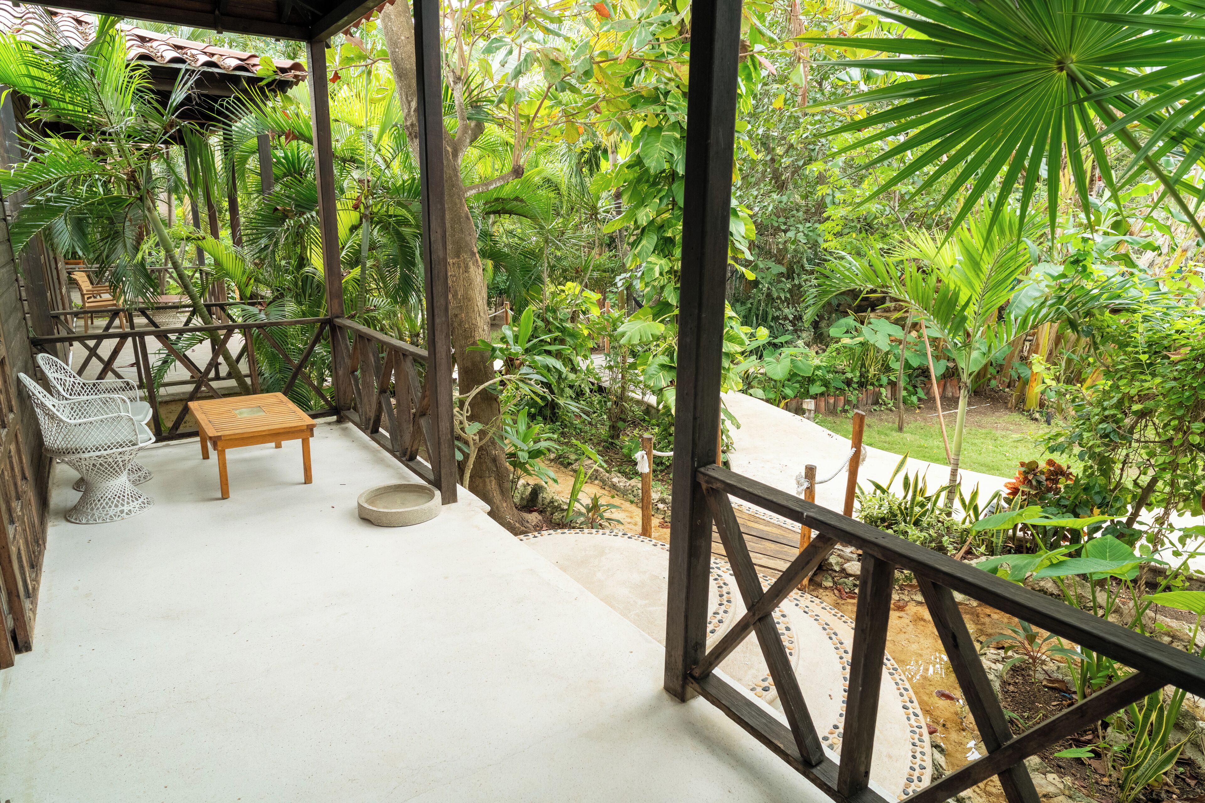 A covered outdoor area with a wooden table and chairs, surrounded by lush tropical plants and a stone pathway.