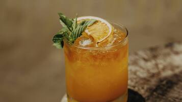A glass of amber-colored beverage with a slice of orange and a mint leaf garnish, placed on a wooden surface.