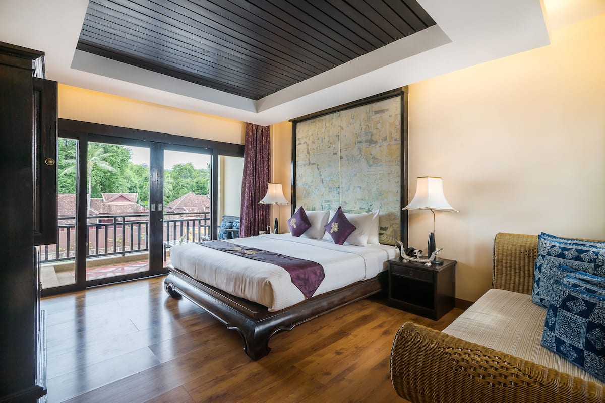 A spacious bedroom with a large bed, a nightstand, a wicker chair, and a balcony with a view of greenery.