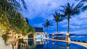 Una piscina infinita tranquila con vistas al océano, palmeras y un edificio con zona de bar.