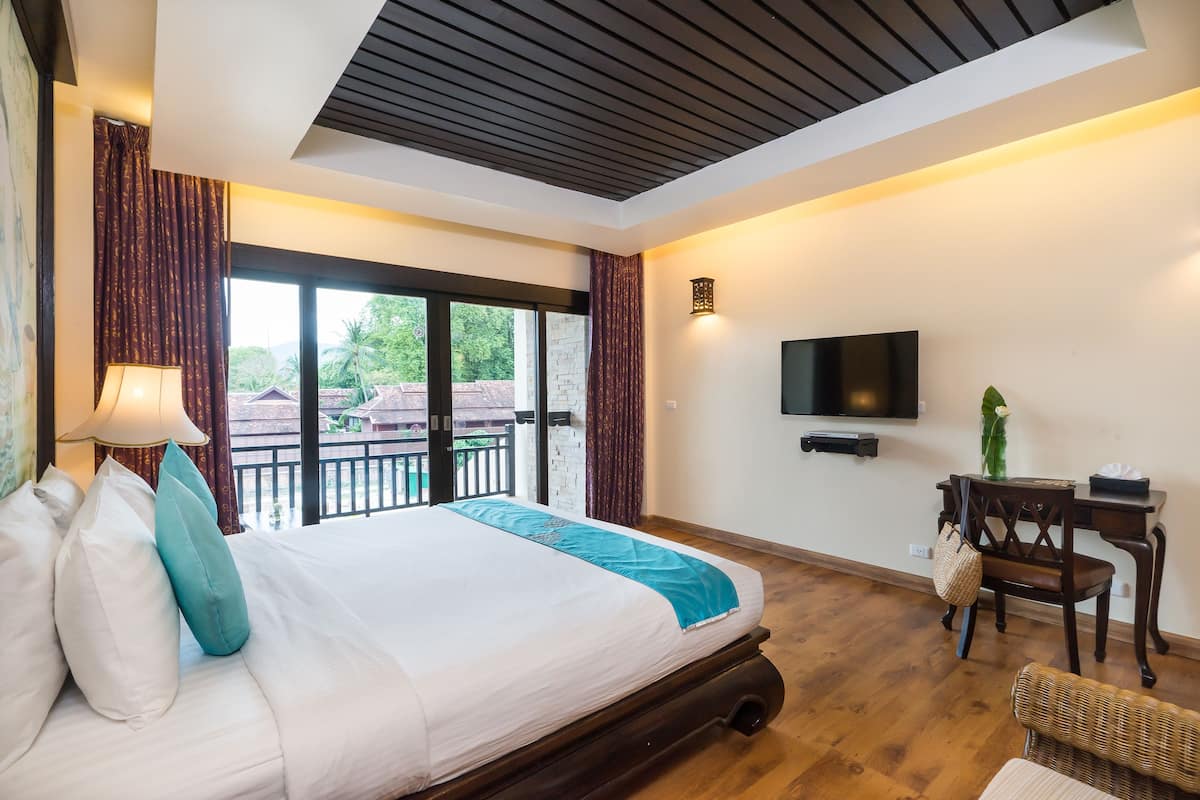 A hotel room with a large bed, a TV mounted on the wall, a desk with a chair, and a balcony with a view of greenery.