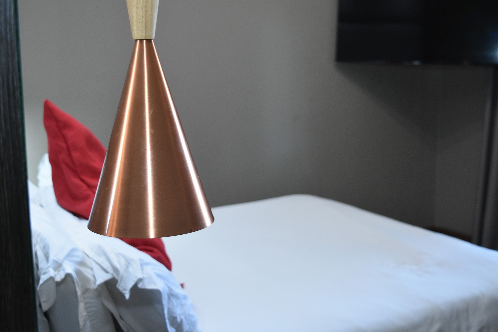 Een eenpersoonsbed met een wit laken en een rood kussen, een koperkleurige hanglamp, en een donkere muur met een flatscreen televisie erboven.
