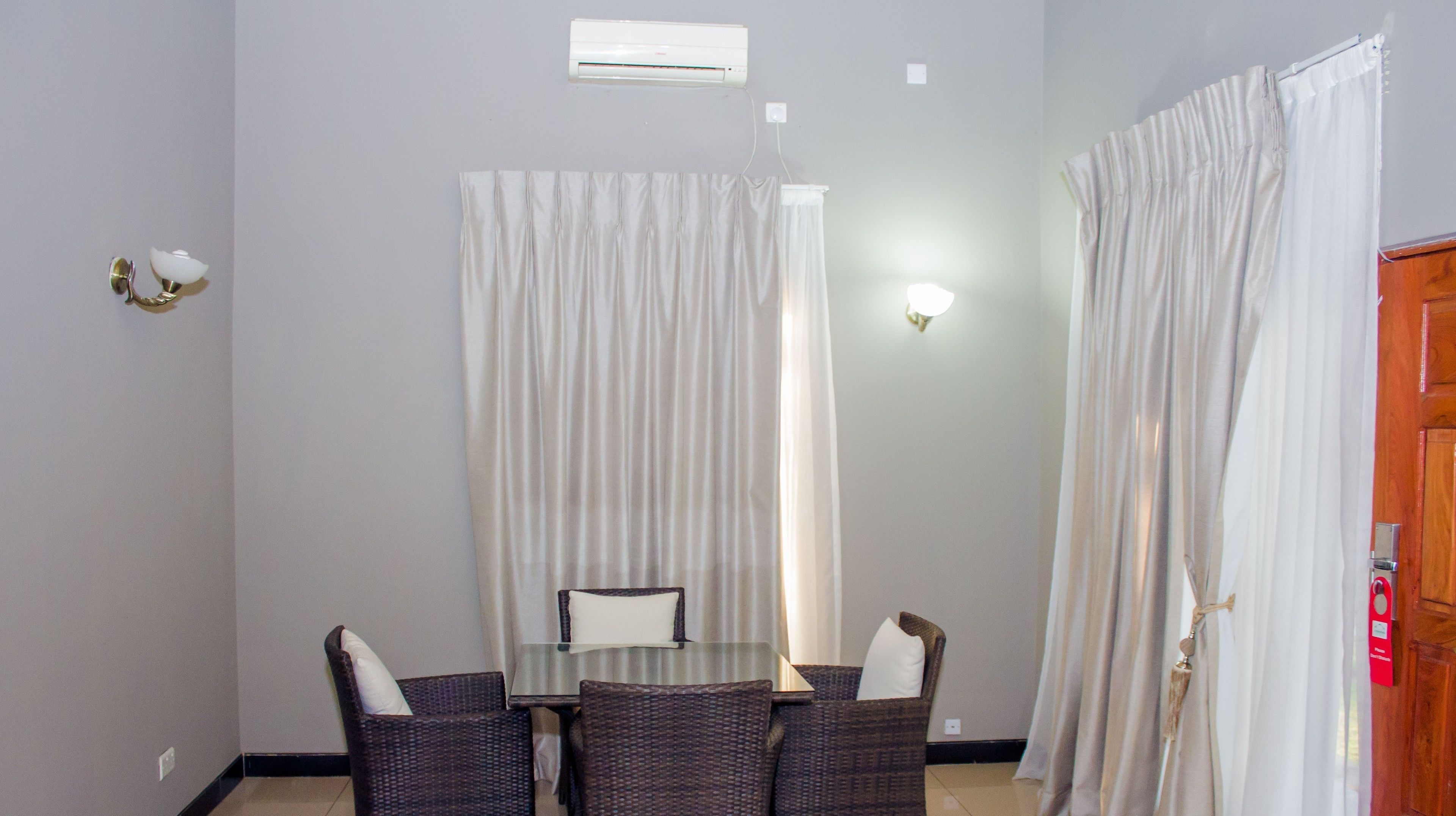 Une pièce comprenant une table à manger, quatre chaises, des rideaux blancs, un luminaire fixé au mur et une porte ornée d’une étiquette rouge.