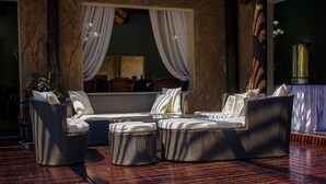 A sunlit outdoor seating area with wicker furniture and a view of the interior through a window with white curtains. - Chrismar Hotel Livingstone (Livingstone)