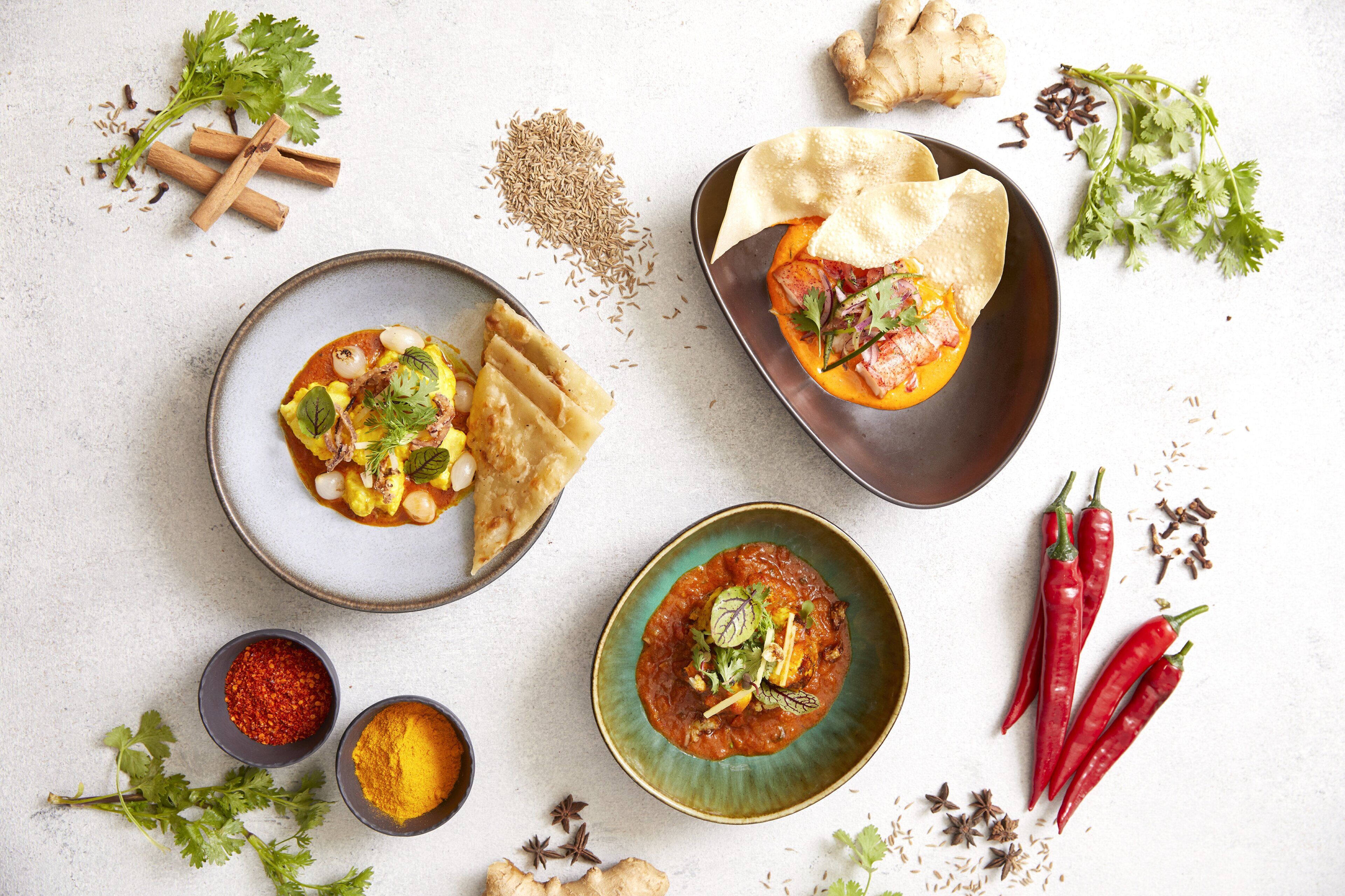 Ein Tisch mit verschiedenen Speisen und Gewürzen, darunter eine Curry-Schüssel, Fladenbrote und ein Teller mit gefülltem Gemüse.