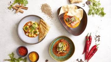 Ein Tisch mit verschiedenen Speisen und Gewürzen, darunter eine Curry-Schüssel, Fladenbrote und ein Teller mit gefülltem Gemüse.