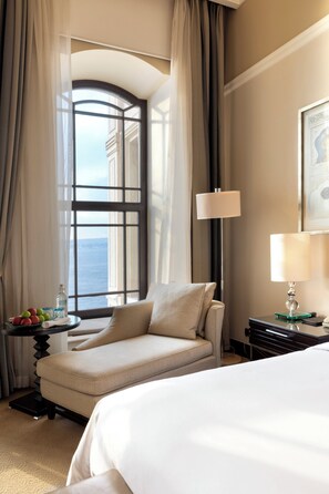 A hotel room with a large bed, a chair, a table with fruits and a bottle, and a view of the sea through a window.