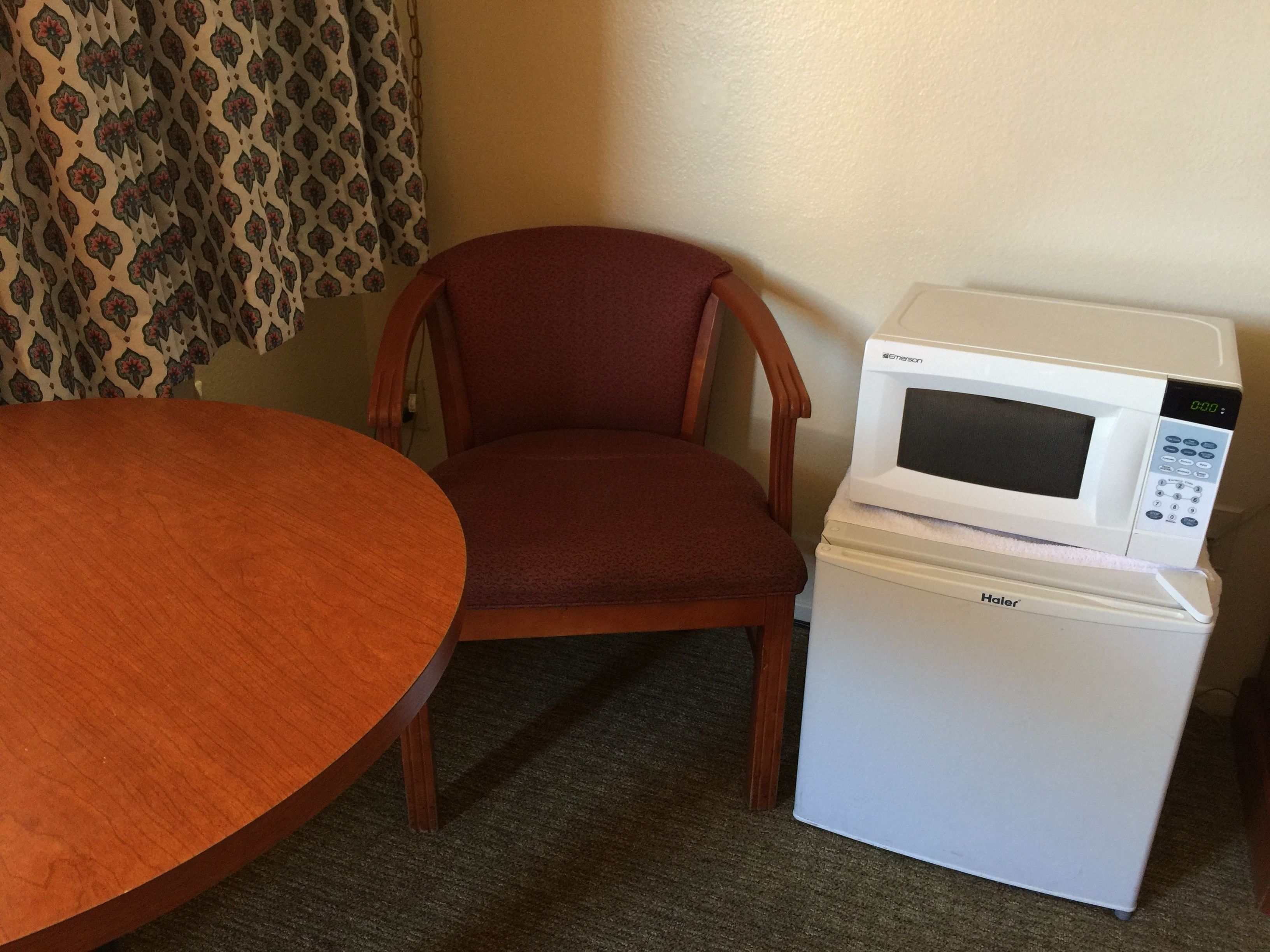 A compact kitchenette with a microwave on a Haier refrigerator, a wooden dining table, and a patterned curtain.