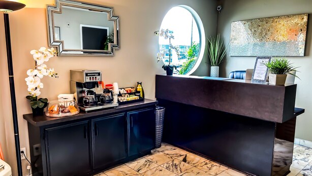 Un espace d’accueil avec un comptoir sombre, un coin café, un grand miroir et une plante en pot.