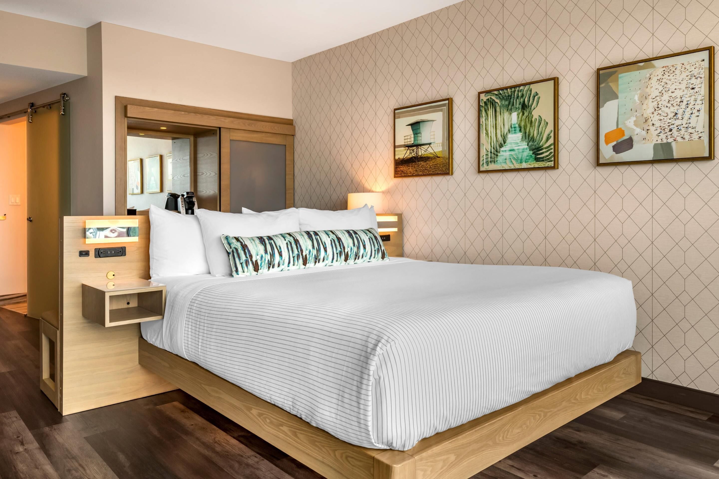 A modern hotel room with a large bed, wooden headboard, bedside tables, and artwork on the walls.