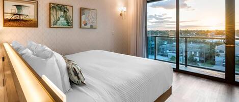 A modern hotel room with a large bed, a wooden nightstand, and a view of the city through floor-to-ceiling windows.