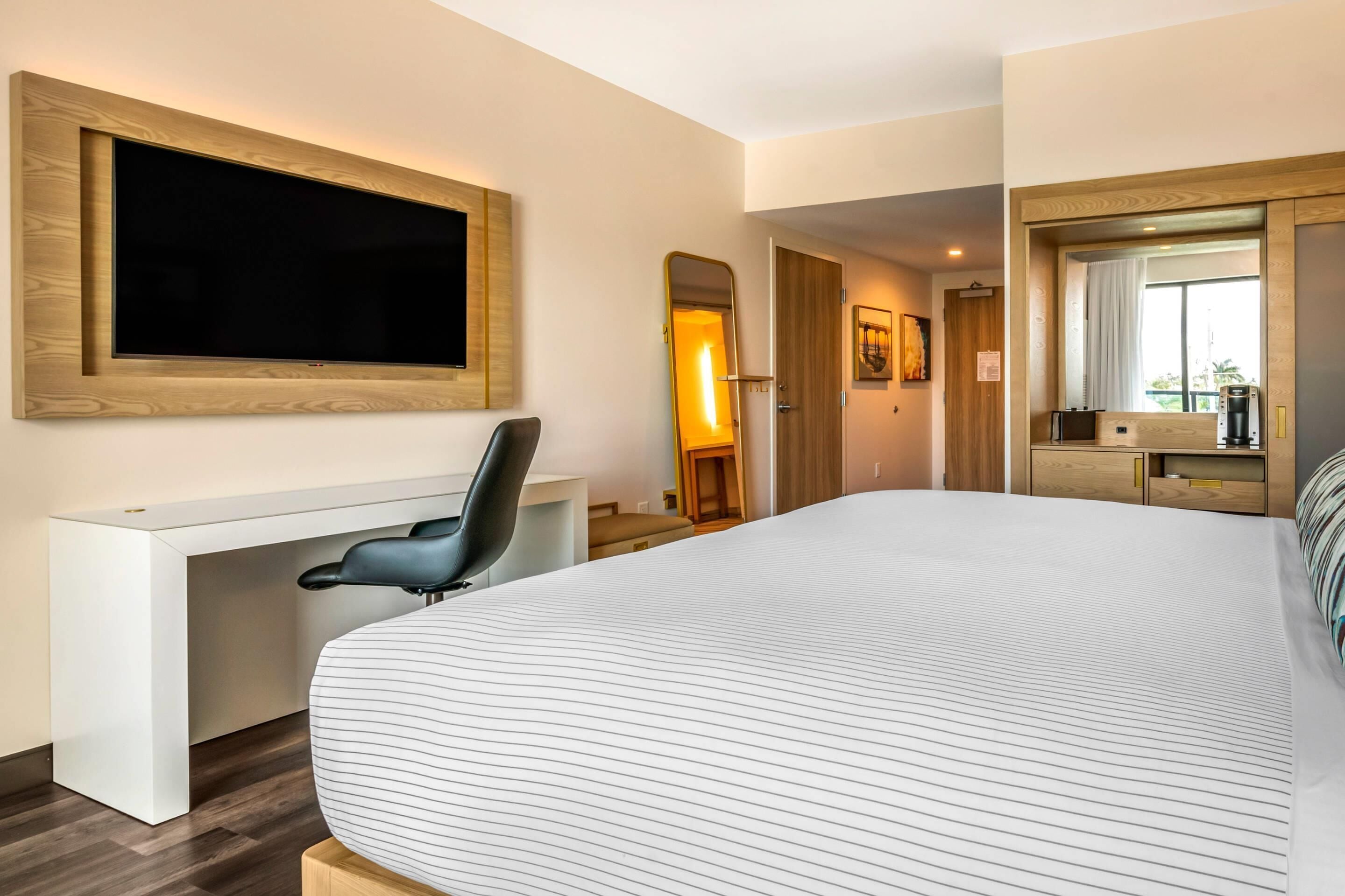 Ein modernes Hotelzimmer mit einem großen Bett, einem Schreibtisch mit Stuhl, einem an der Wand montierten Fernseher und einem Spiegel über dem Bett.