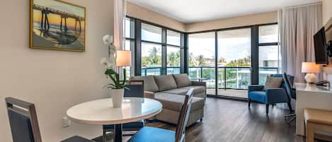 A modern hotel room with a round dining table, two sofas, a TV, and large windows offering a view of palm trees and a bridge.