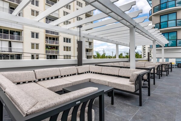 A rooftop lounge area with a long, L-shaped sofa and multiple tables, surrounded by a white pergola structure.