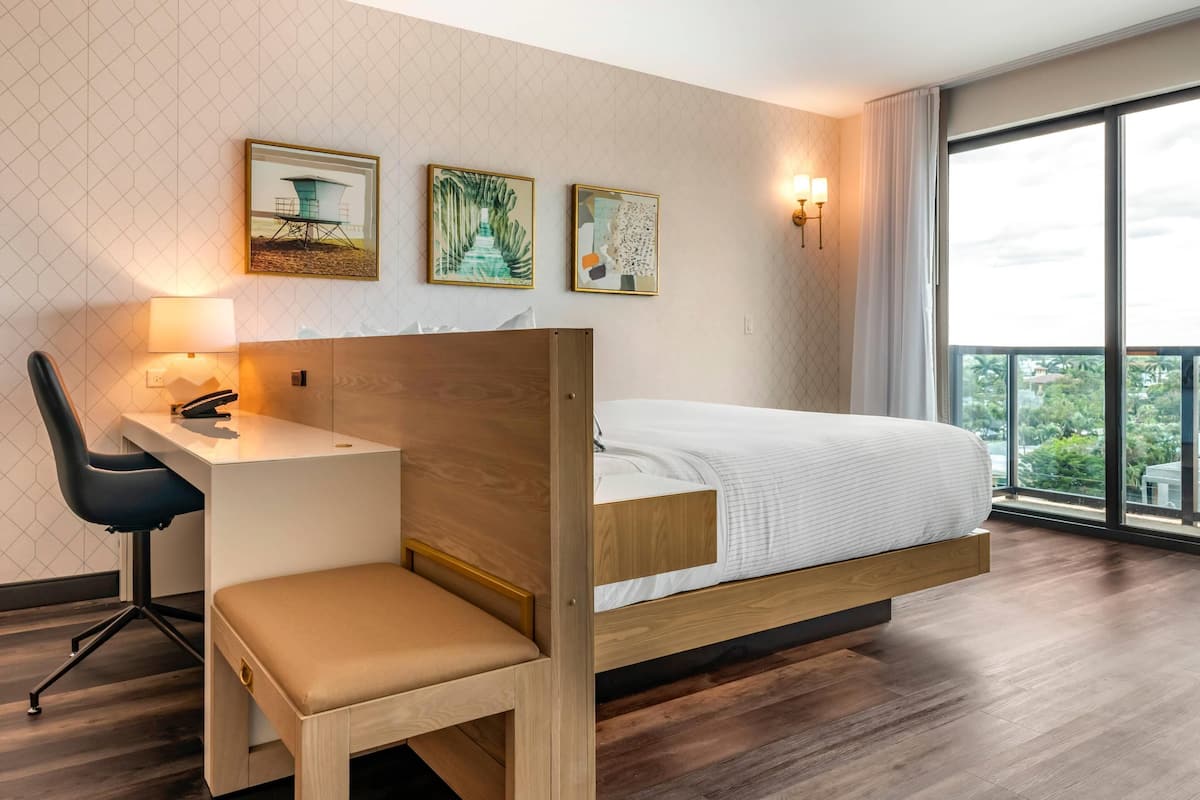 Habitación de hotel moderna con cama de madera, un escritorio y una silla. Hay tres cuadros en la pared y un ventanal con vistas a zonas verdes.