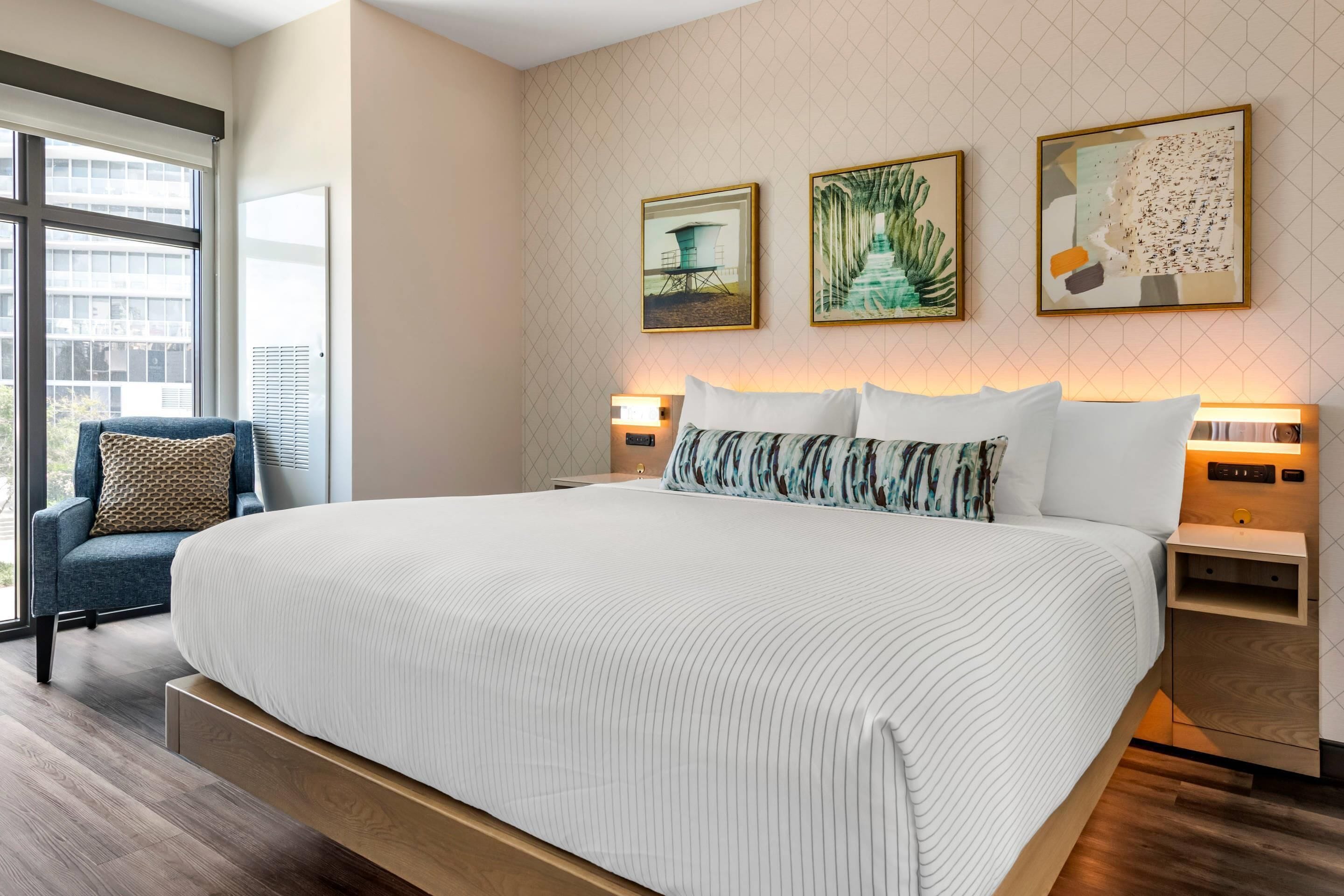 Moderni hotellihuone, jossa on suuri sänky, tuoli ja taideteos seinällä.