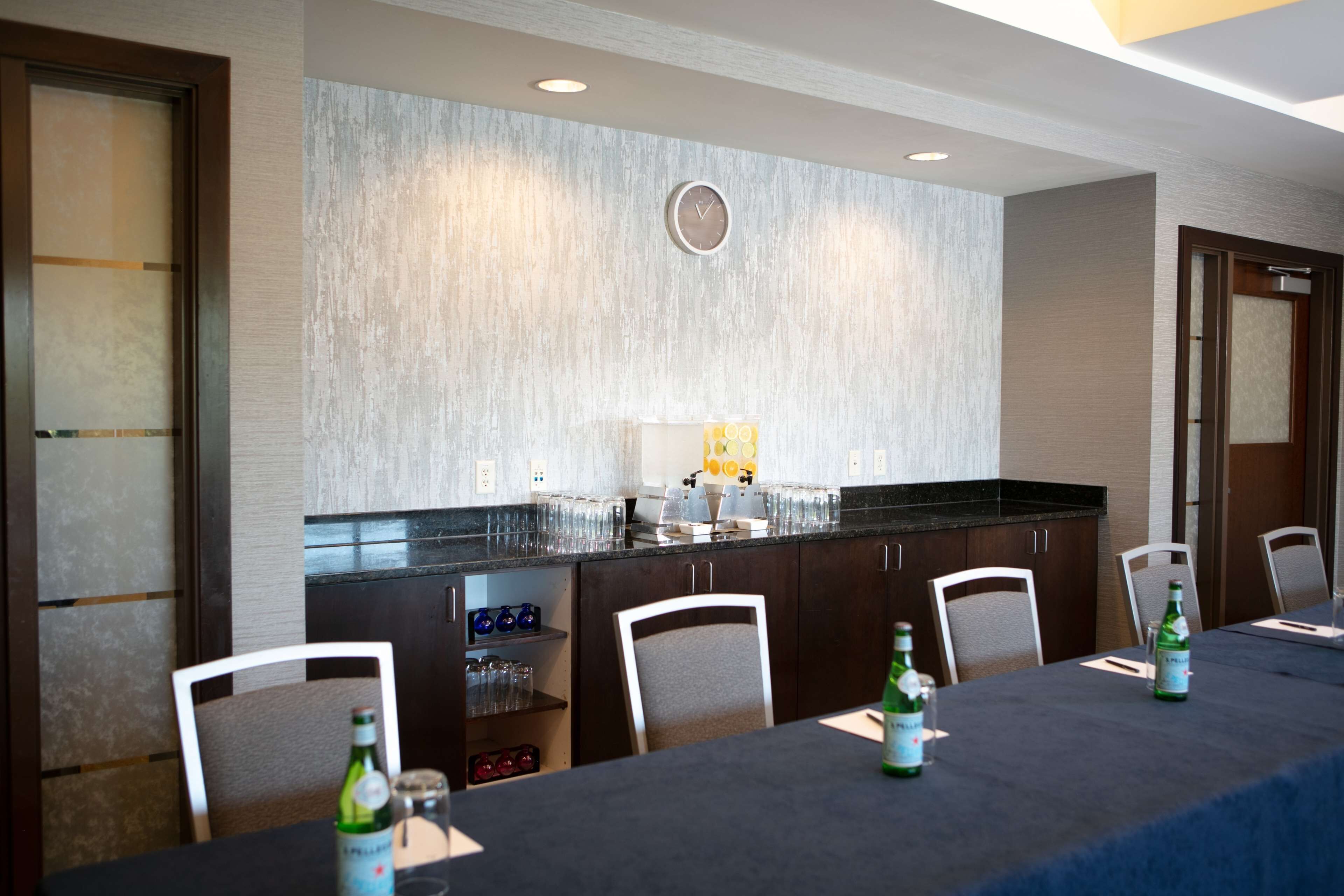 A conference room with a long table, chairs, a clock on the wall, and a kitchen area with a refrigerator and sink.