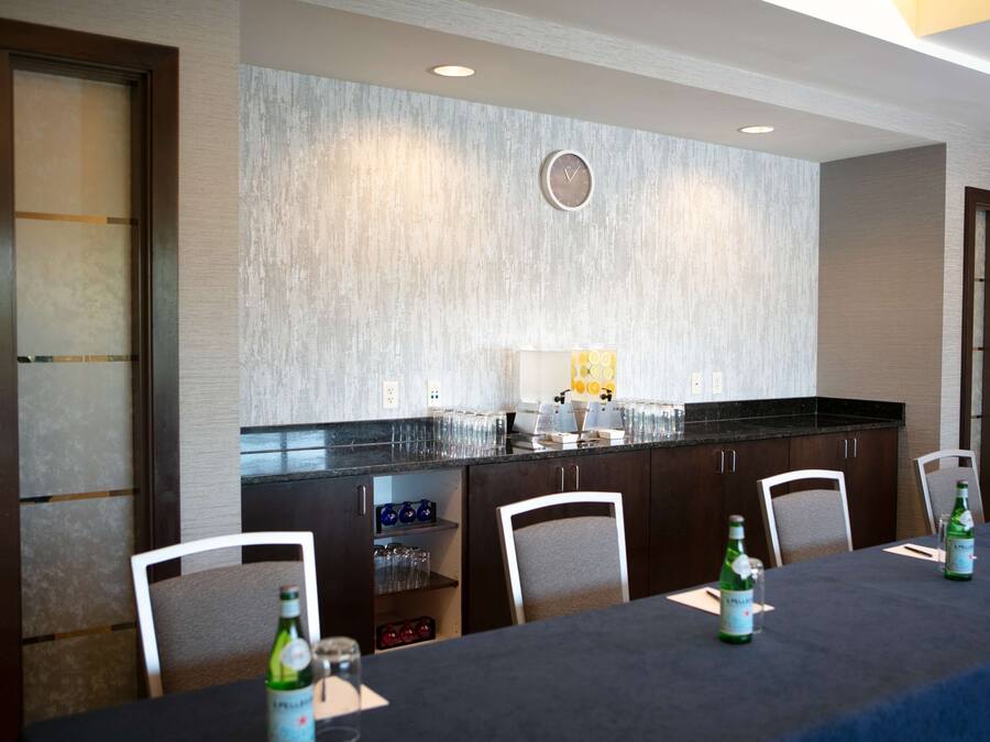 A conference room with a long table, chairs, a clock on the wall, and a kitchen area with a refrigerator and sink.