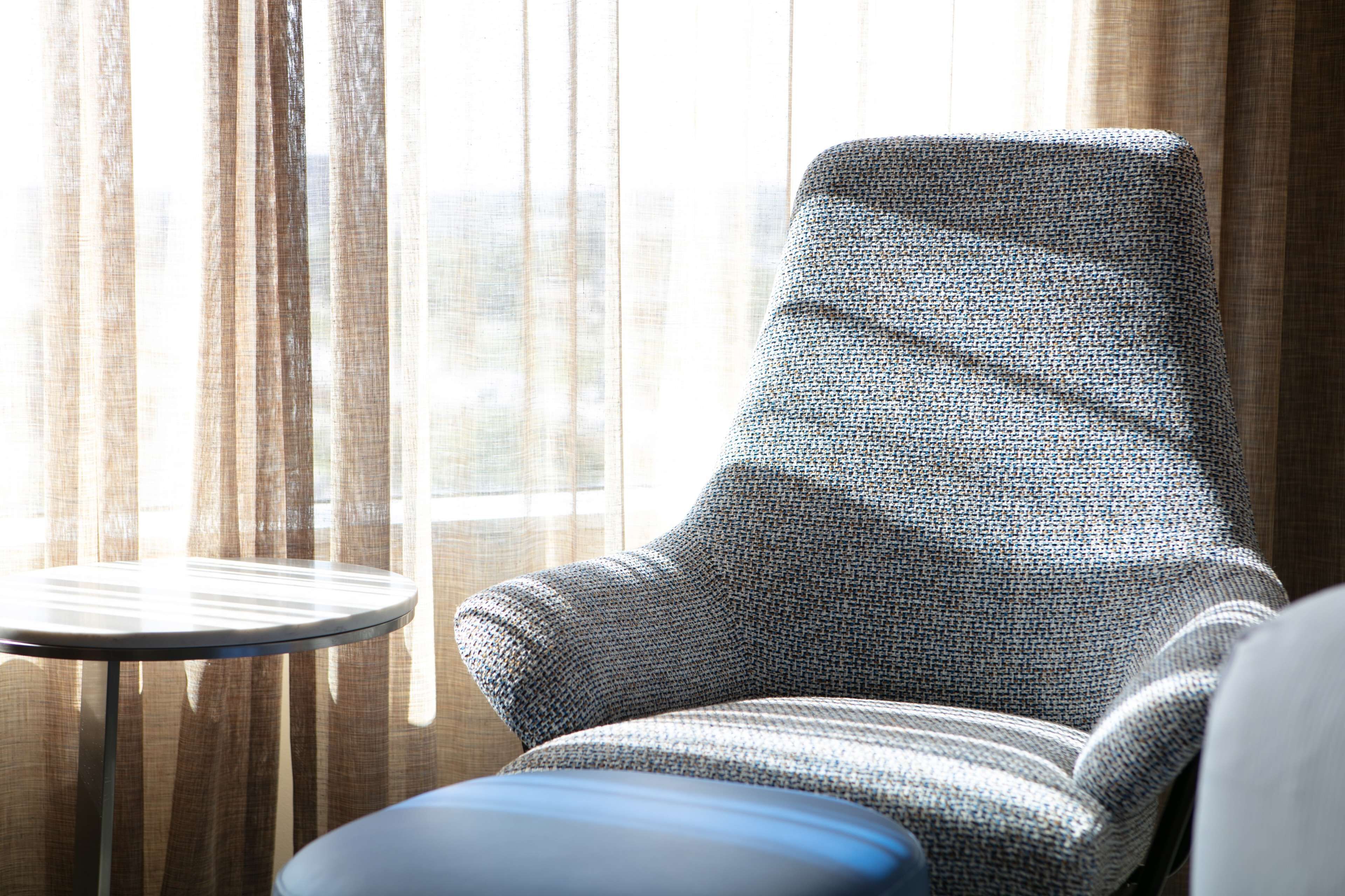 Una habitación con un sillón estampado, una mesita redonda pequeña y cortinas traslúcidas.