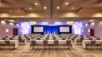A large conference room with rows of tables and chairs, a stage with a blue curtain, and a large screen.