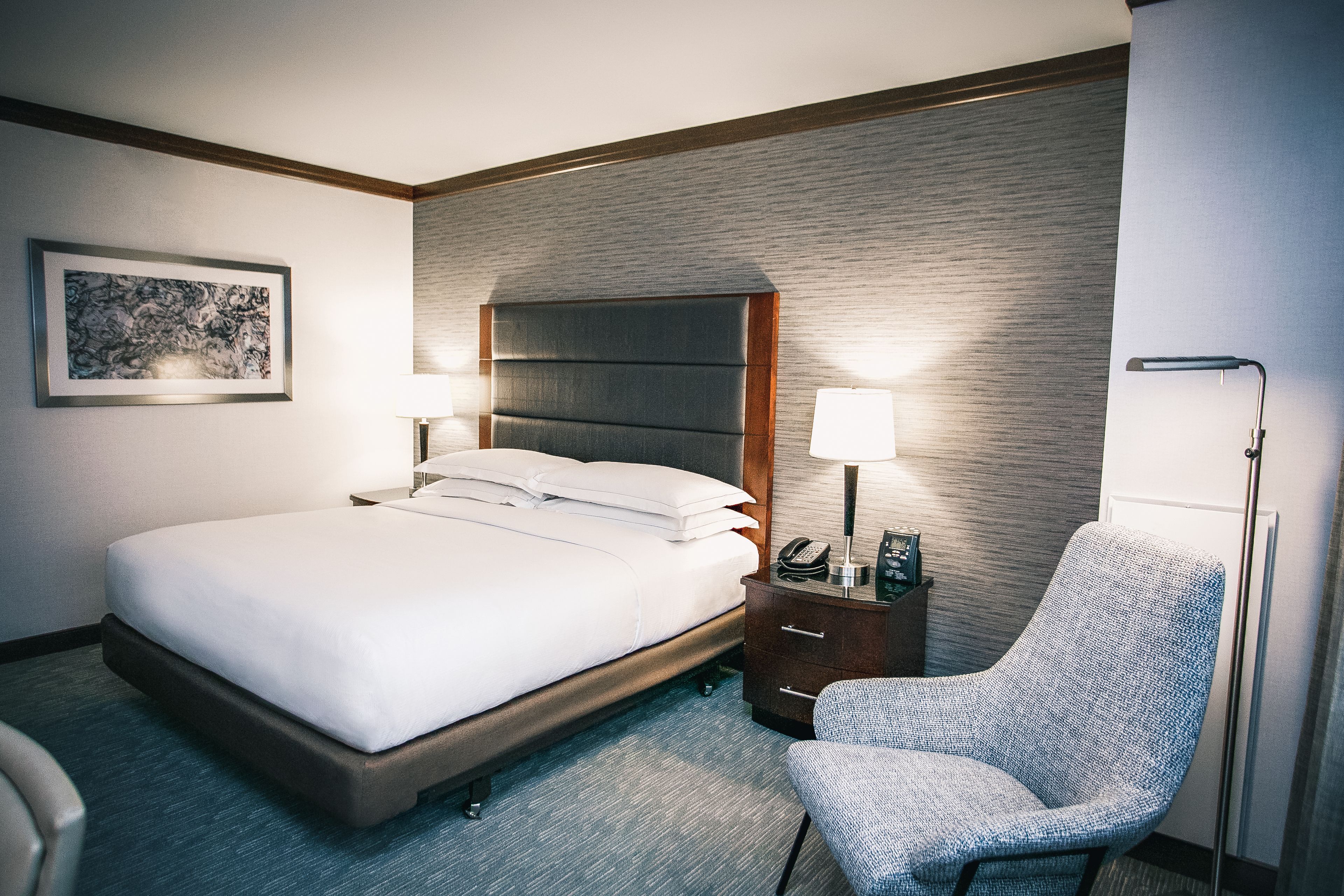 Quarto de hotel com uma cama grande, mesas de cabeceira, uma cadeira e um quadro na parede.
