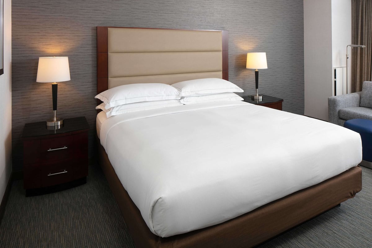 A neatly made bed with white linens, two bedside tables with lamps, a dark wooden headboard, and a grey armchair.