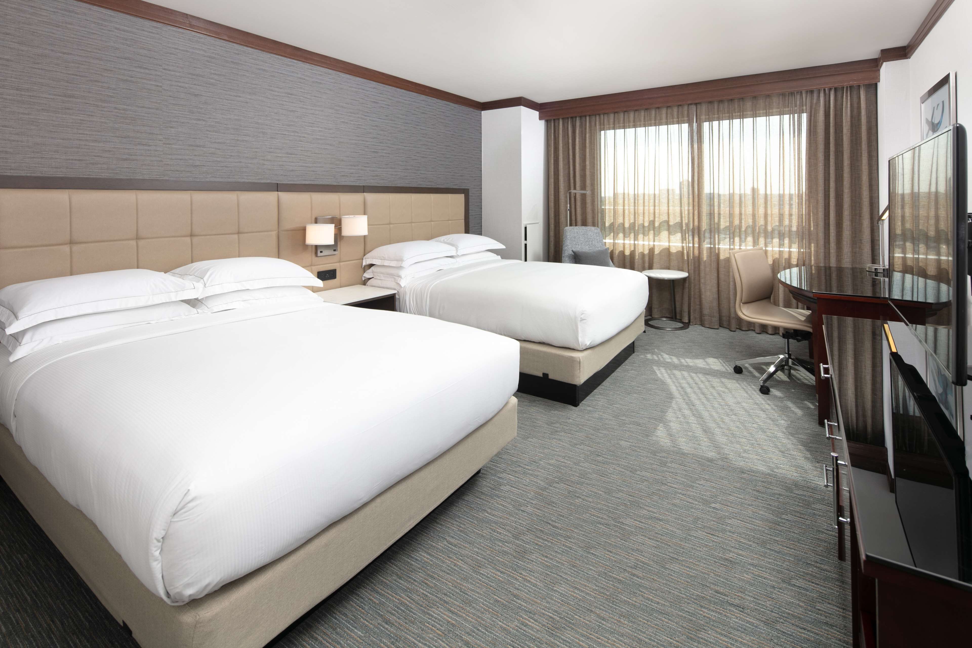 A hotel room with two beds, a desk, a chair, a TV, and a large window with curtains.