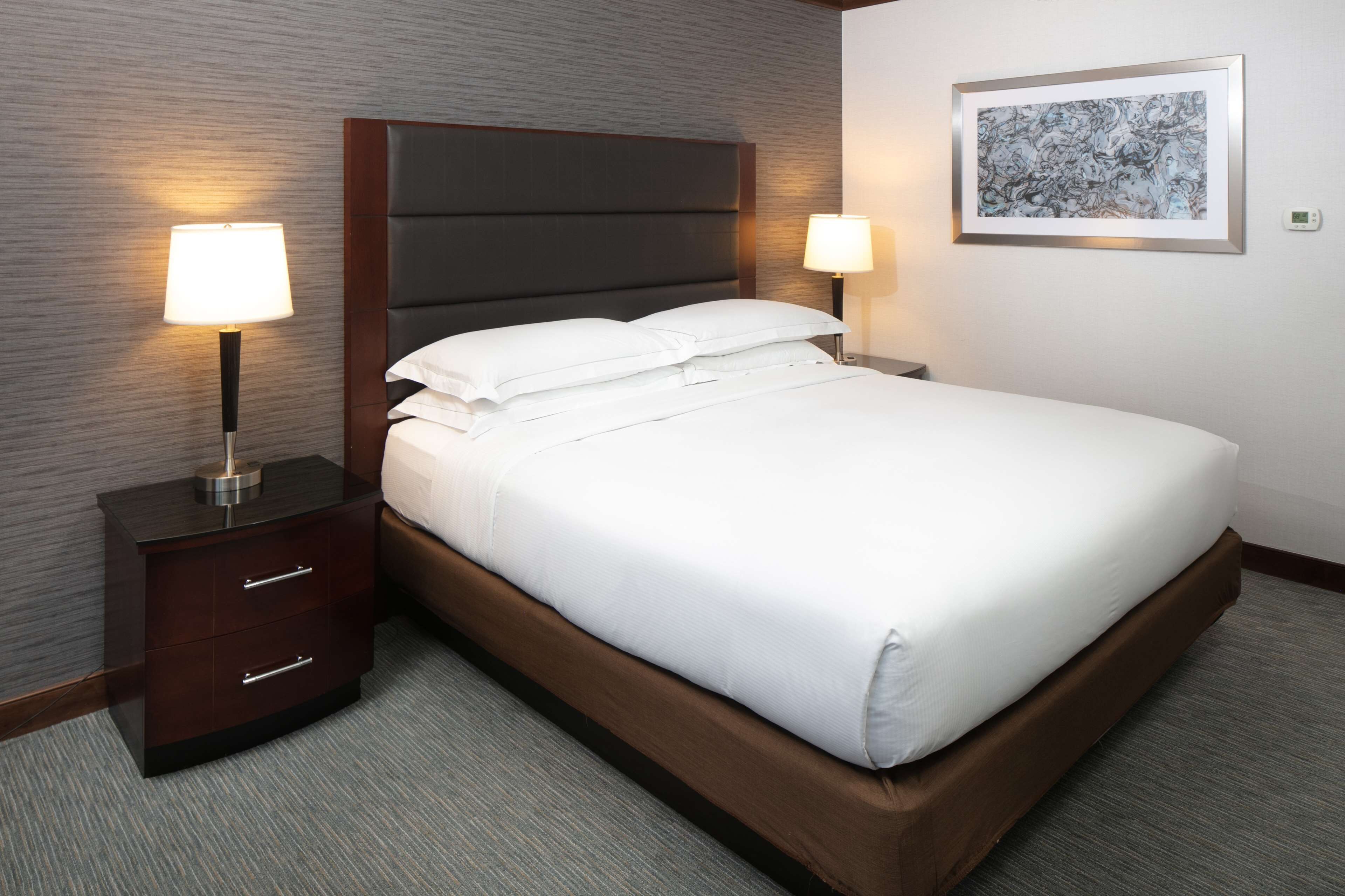 Una cama bien hecha con sábanas blancas, un cabecero oscuro y dos lámparas de noche sobre mesitas de madera.