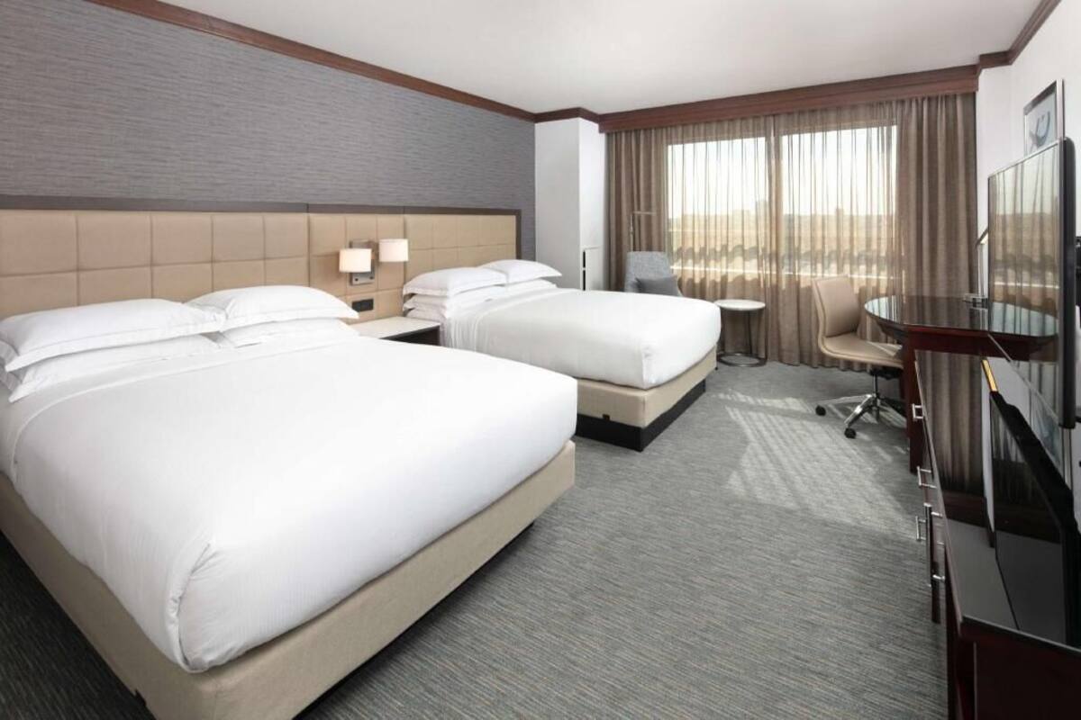 A hotel room with two beds, a desk, a chair, a TV, and a large window with curtains.