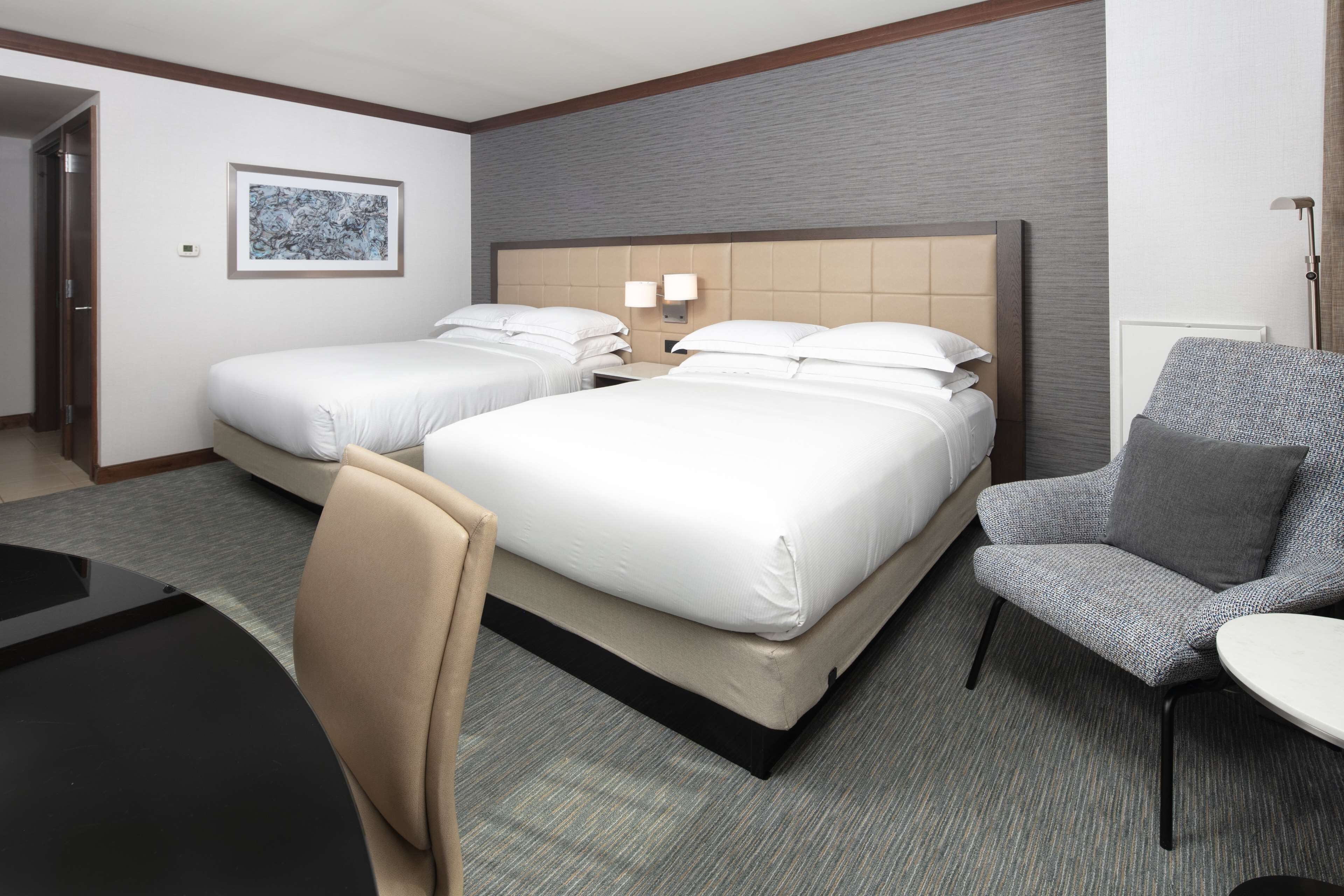 Quarto de hotel com duas camas, uma escrivaninha, uma cadeira e uma luminária.
