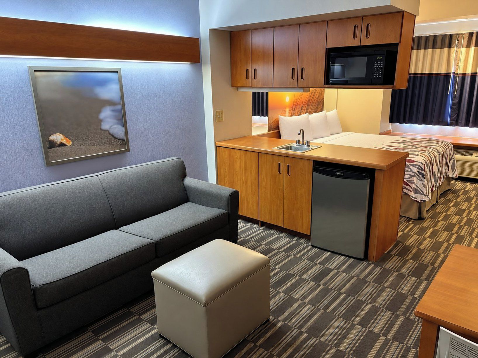 Habitación de hotel compacta con sofá, un pequeño reposapiés, kitchenette con microondas y cama con manta a rayas.