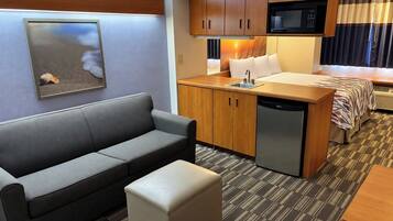 A compact hotel room with a sofa, a small ottoman, a kitchenette with a microwave, and a bed with a striped blanket.