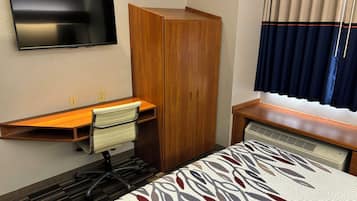 Una habitación de hotel con una cama, un escritorio con silla, un armario, un televisor montado en la pared y una ventana con cortinas.