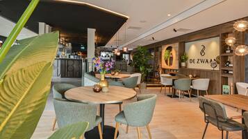 A modern interior with round tables and chairs, a bar area, and a sign reading "DE ZWAAN".