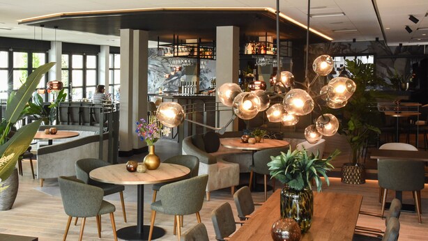 Een modern restaurantinterieur met ronde tafels, grijze gestoffeerde stoelen en een centraal hanglamp met bolvormige lampen.