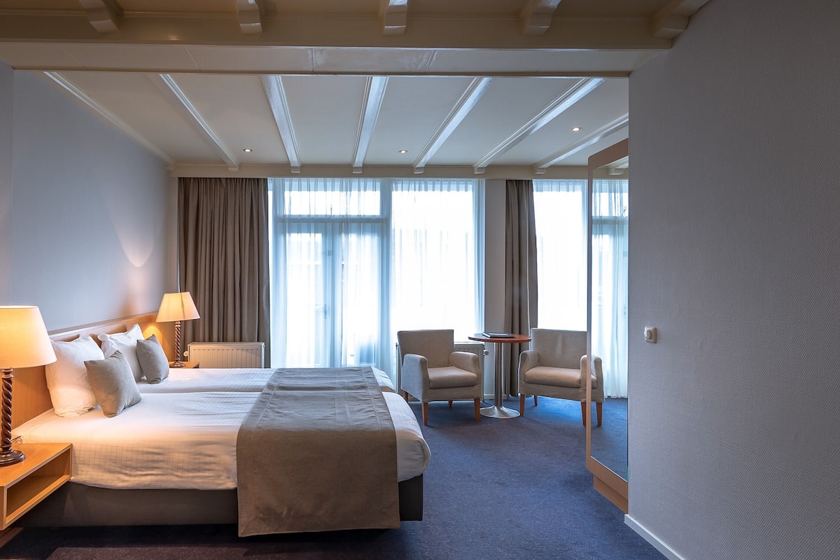 Een hotelkamer met twee bedden, een zithoek met stoelen, een kleine tafel en grote ramen met gordijnen.