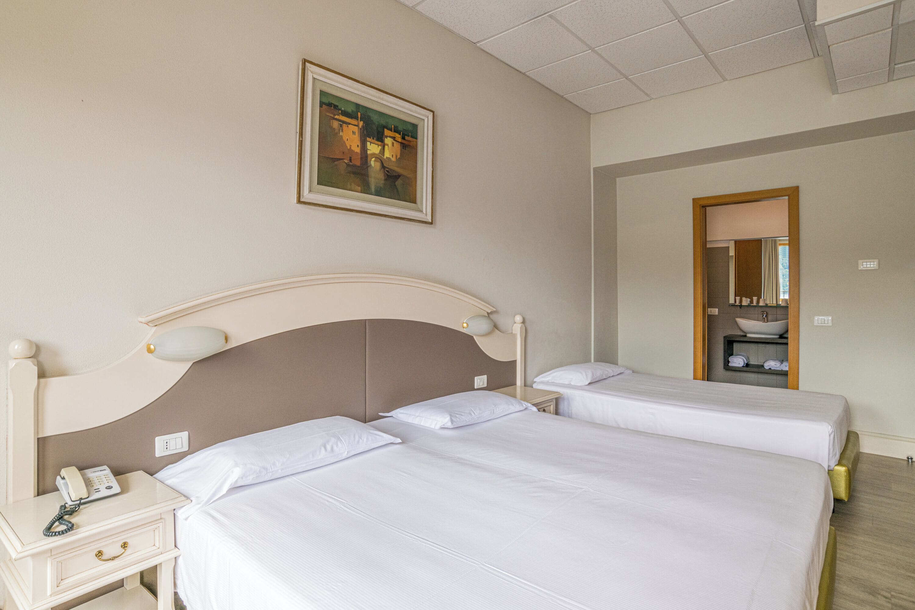 A hotel room with two beds, a bedside table with a telephone, a painting on the wall, and a bathroom visible through an open door.