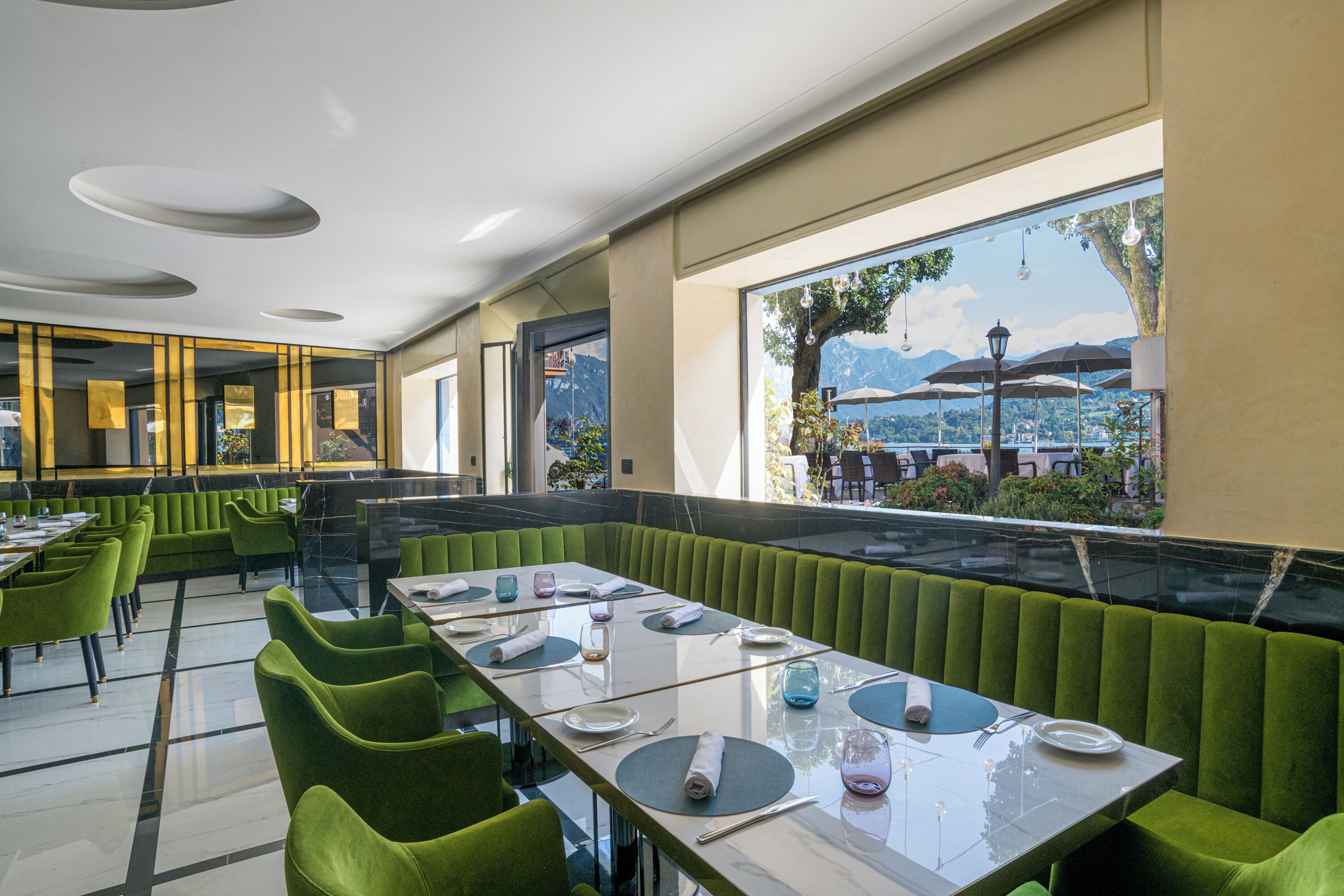 Un restaurante con asientos y mesas tapizados en verde, amplias ventanas que ofrecen vistas a montañas y un lago.
