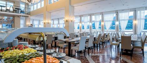 A buffet area with a variety of fruits and vegetables, multiple dining tables with chairs, and a chandelier.